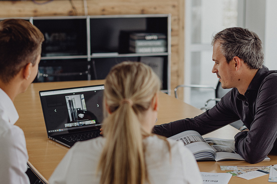 Immobilienberatung mit Laptop und Katalog – Makler zeigt jungem Paar Visualisierung von Wohnung oder Haus
