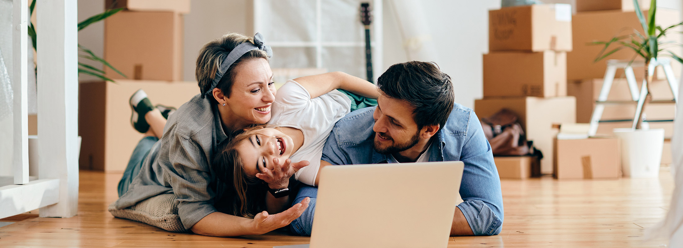 Lachende Familie liegt zwischen Umzugskartons auf dem Boden und schaut gemeinsam auf einen Laptop – neuer Lebensabschnitt