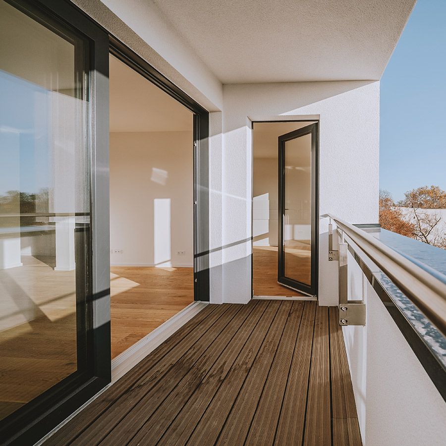 Sonniger Balkon mit Holzbelag und Glasgeländer, Zugang zu hellen Wohnräumen in moderner Neubauwohnung