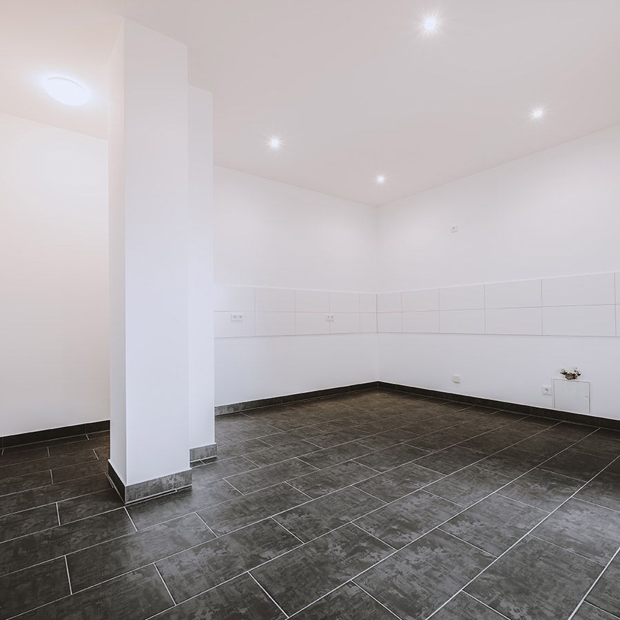 Leerer Küchenraum mit dunklem Fliesenboden, weißen Wänden und Anschlüssen – bereit für Einbauküche