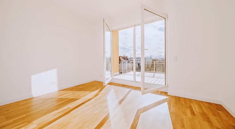 Helles, leeres Wohnzimmer mit Parkettboden und Zugang zum Balkon mit Stadtblick – moderne Neubauwohnung